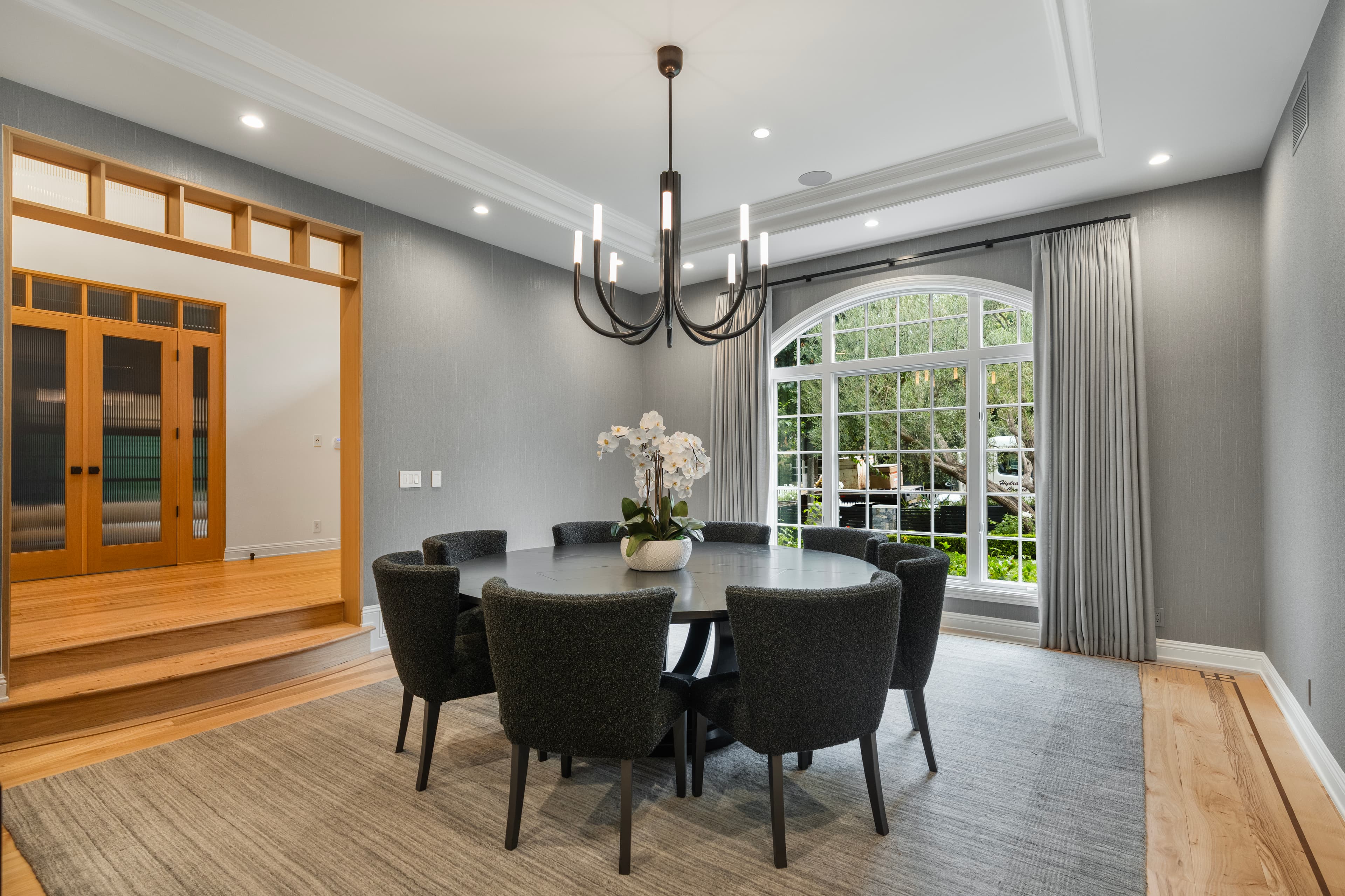 Modern dining room featuring a round black table, grey chairs, and a large arched window.