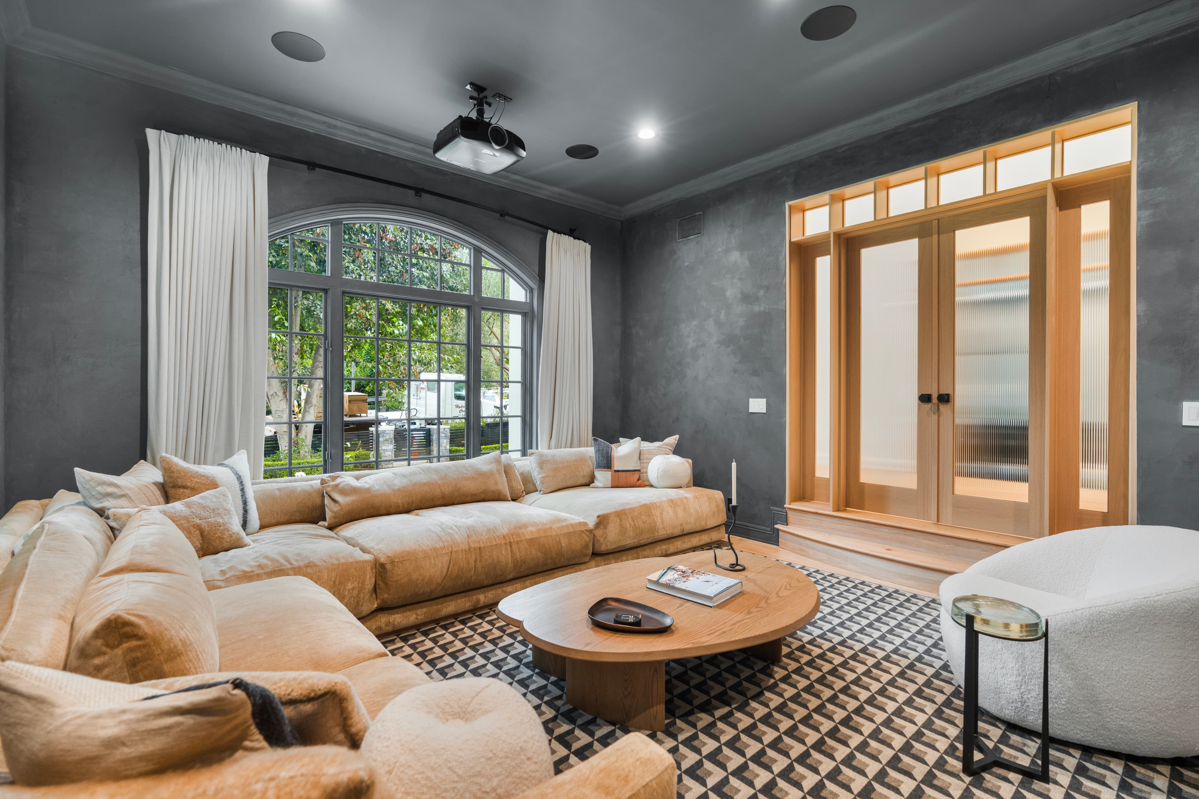Modern media room featuring tan sectional, dark gray walls, arched window, and ceiling projector.
