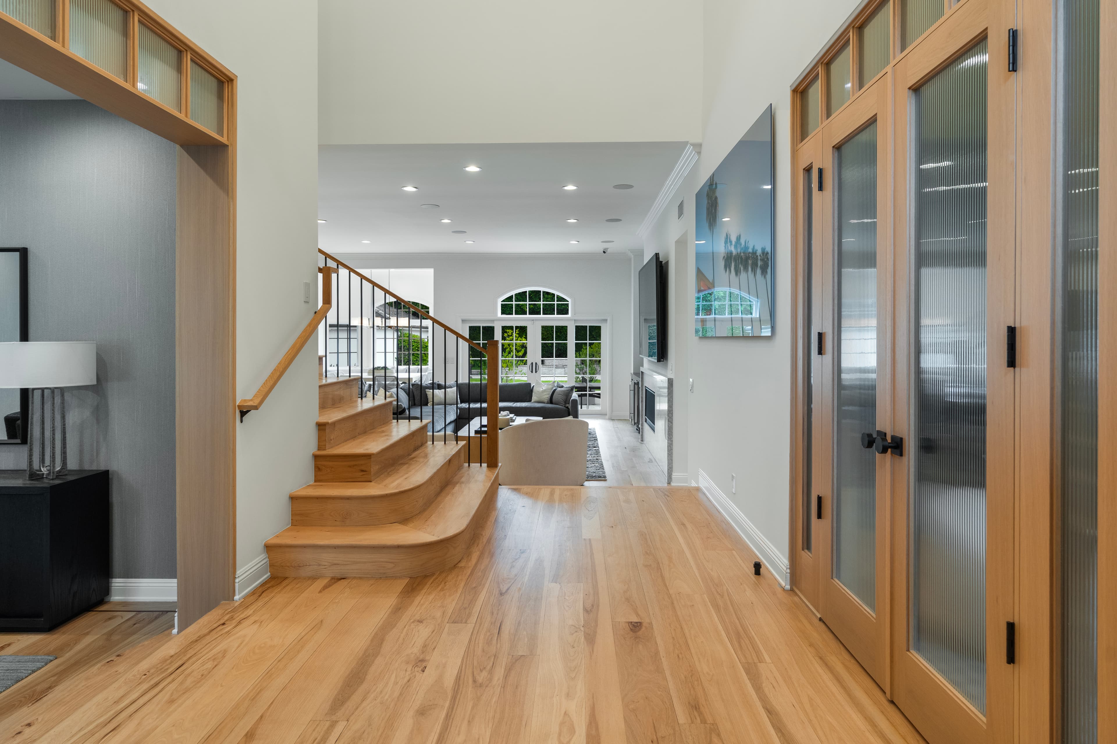 Bright modern hallway with light wood floors, a staircase, and large ribbed glass doors.
