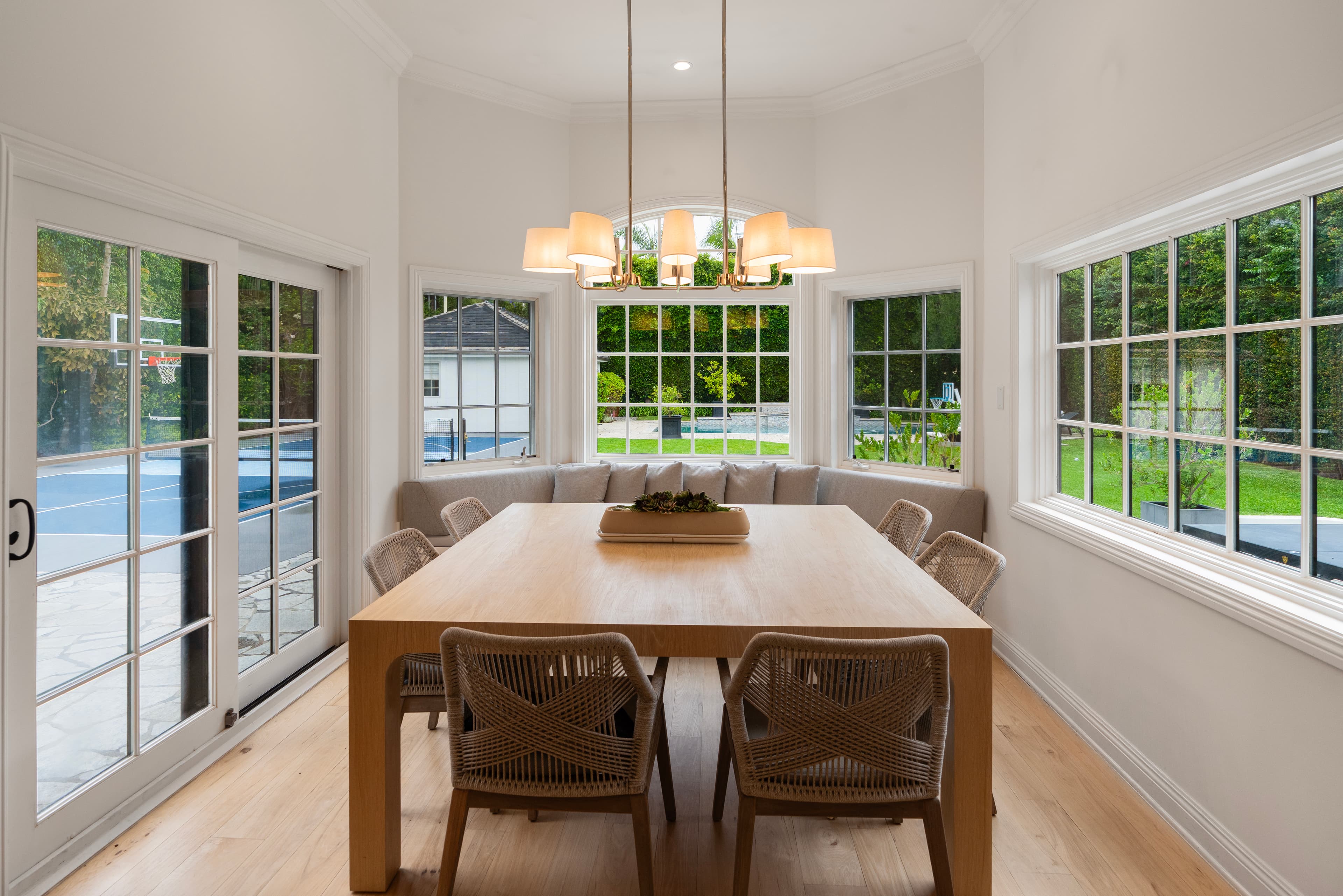 Bright dining room with wooden table, woven chairs, and banquette seating overlooking a backyard.