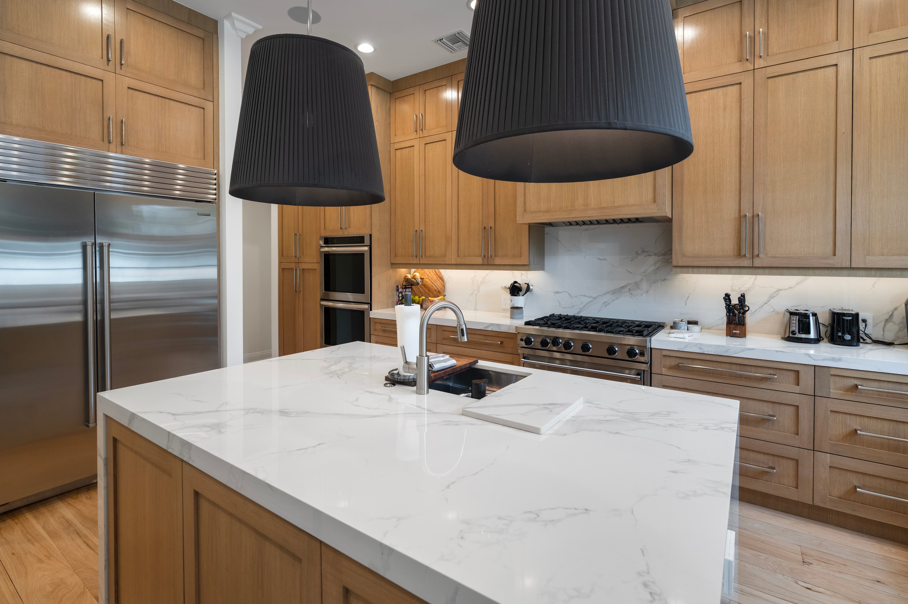 Modern kitchen with light wood cabinets, marble island, stainless steel appliances, and black pendant lights.