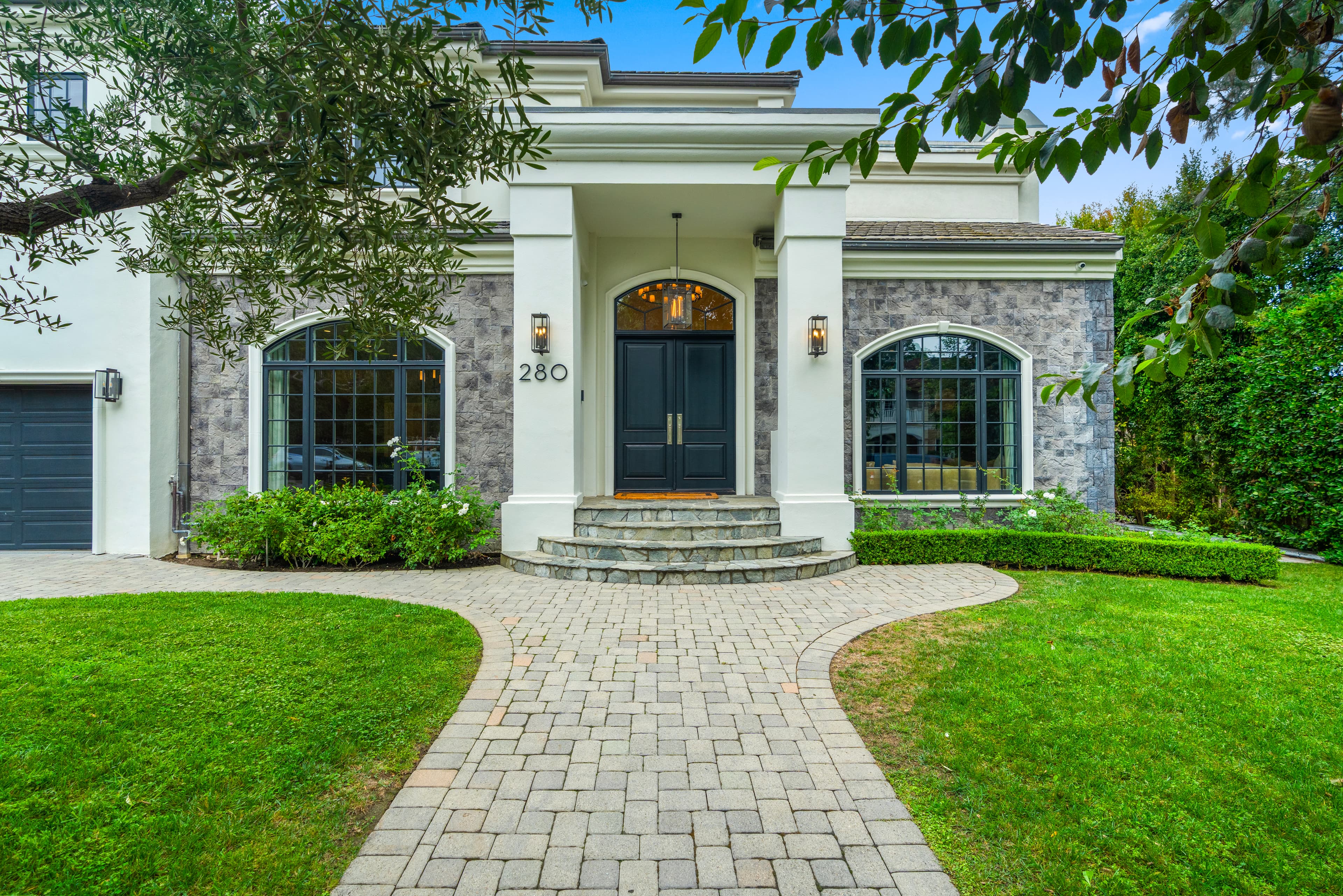 Paved walkway leading to a luxury home entrance with black double doors and stone facade.