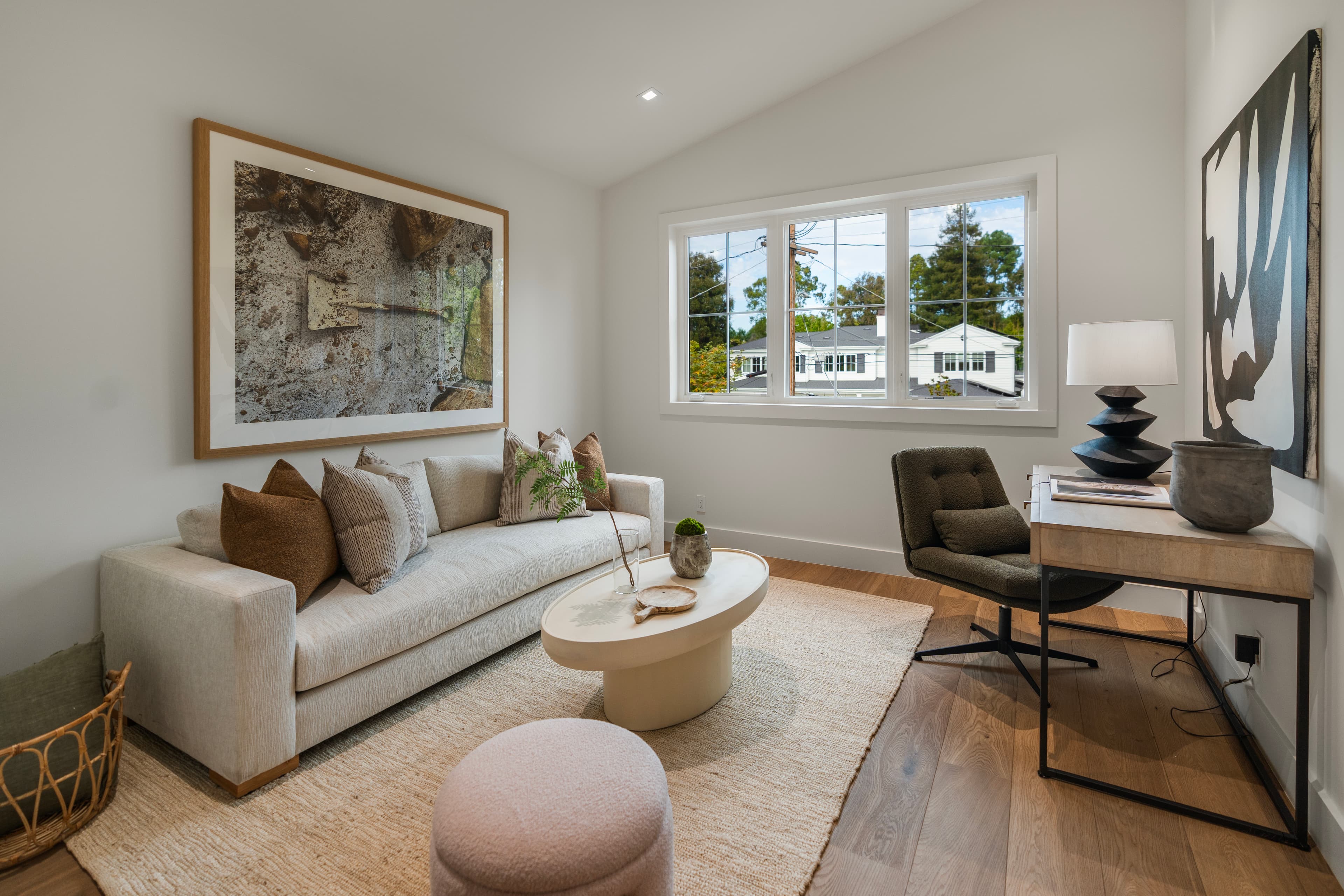 Modern home office with a neutral sofa, oval coffee table, desk, and large framed artwork.