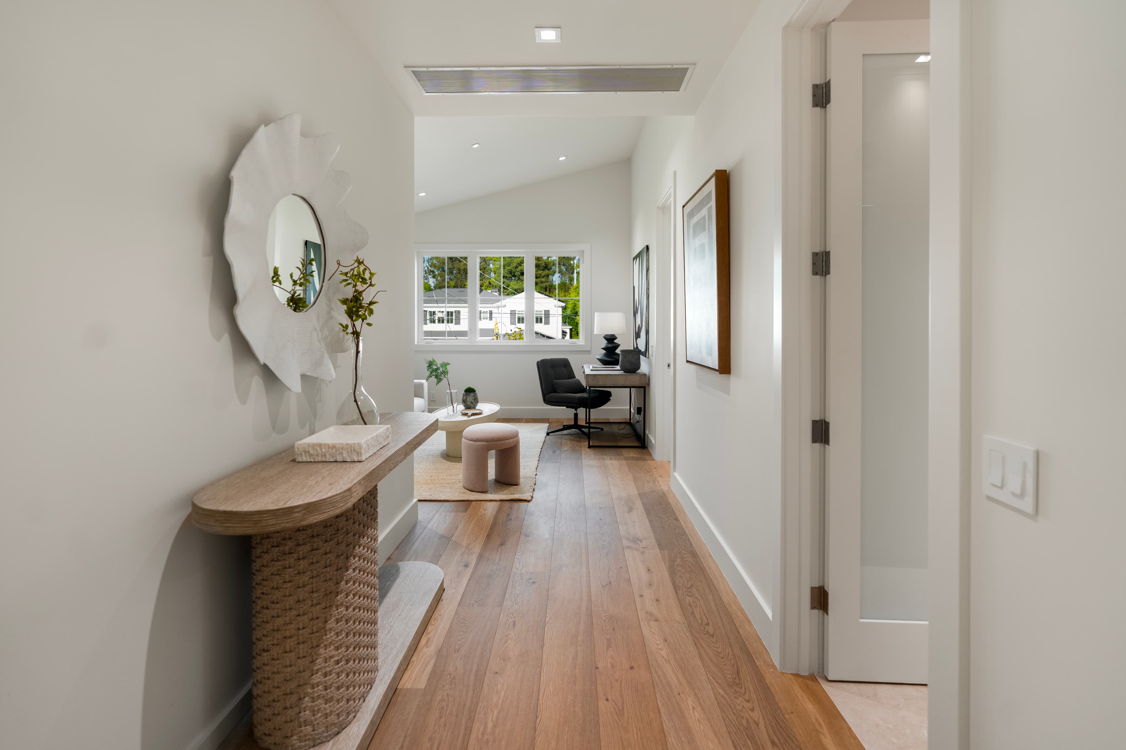 Modern hallway with a wooden console and wavy mirror leading to a bright home office.
