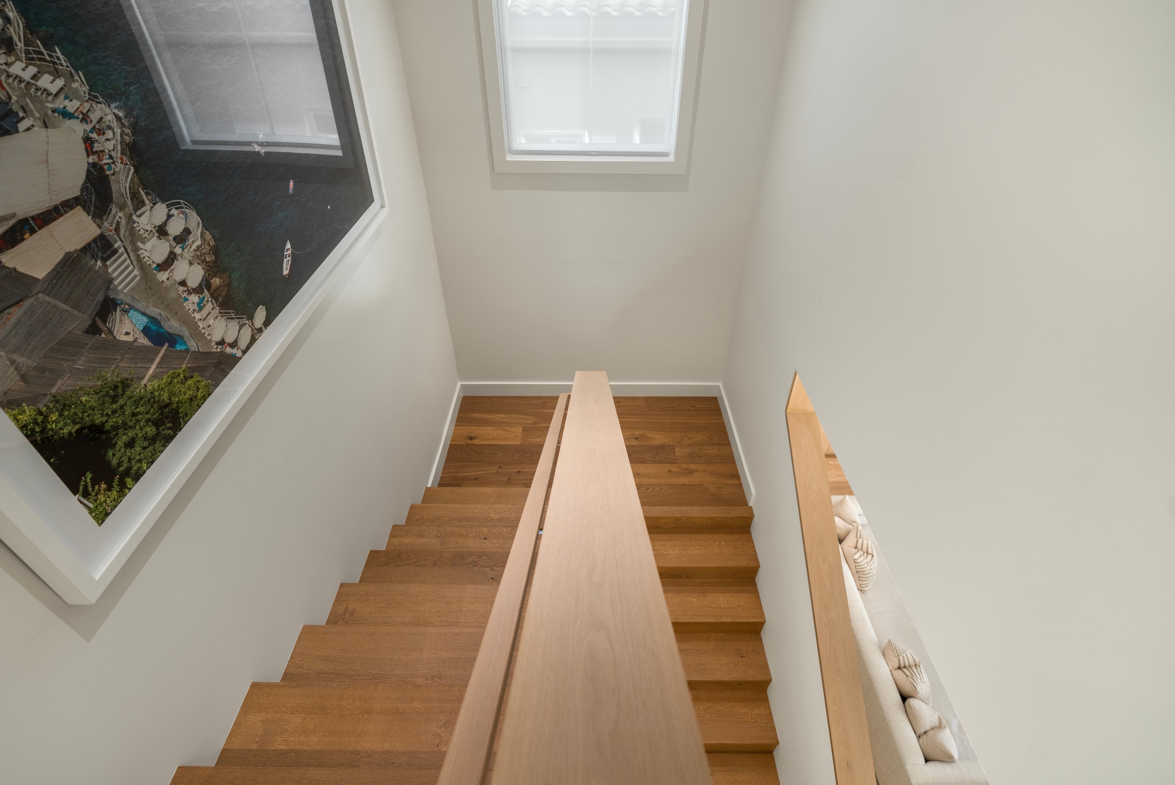 Looking down a wooden staircase with white walls, central handrail, and a large coastal photograph.