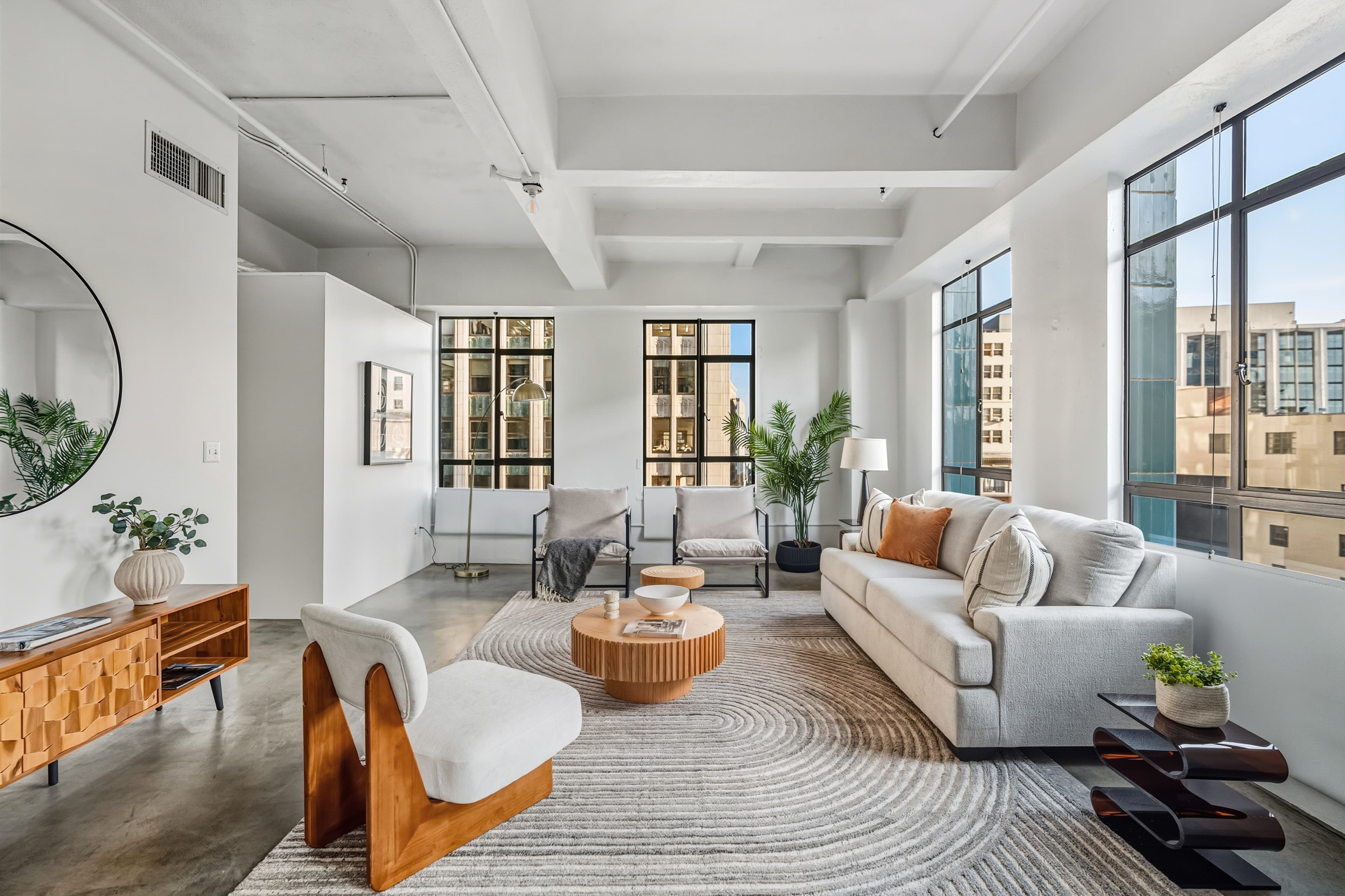 Bright industrial loft living room with white sofa, wooden coffee table, and large city windows.