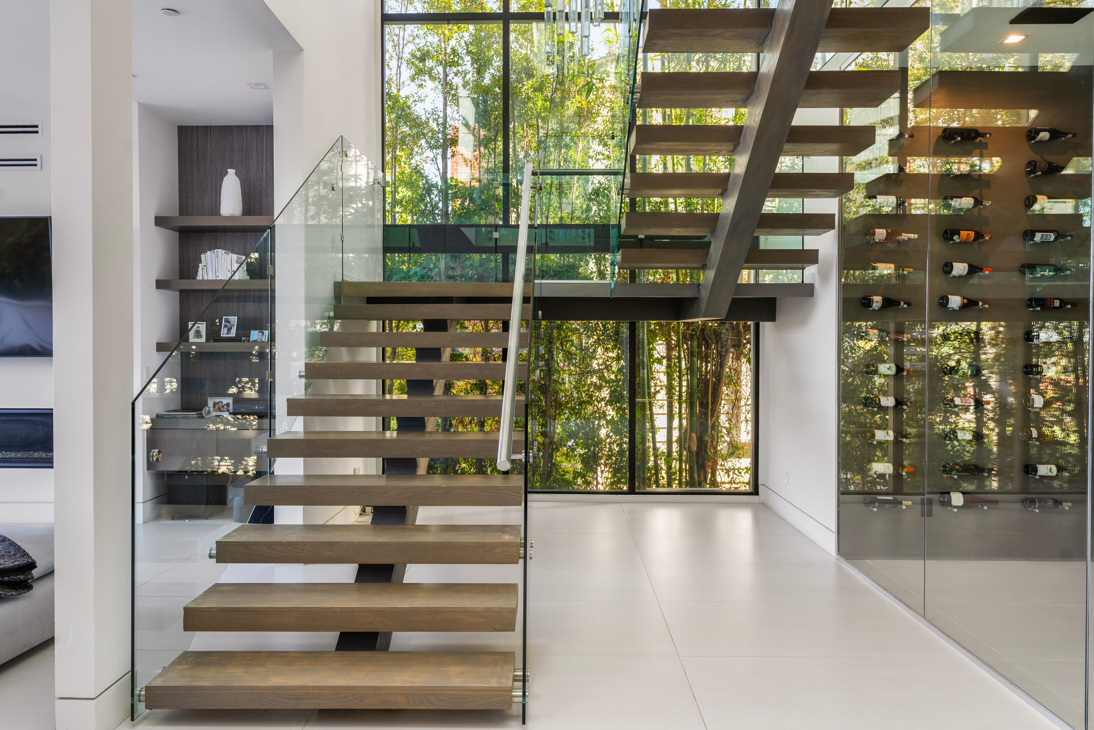 Modern wooden staircase with glass railings, a glass wine cellar, and lush garden views.