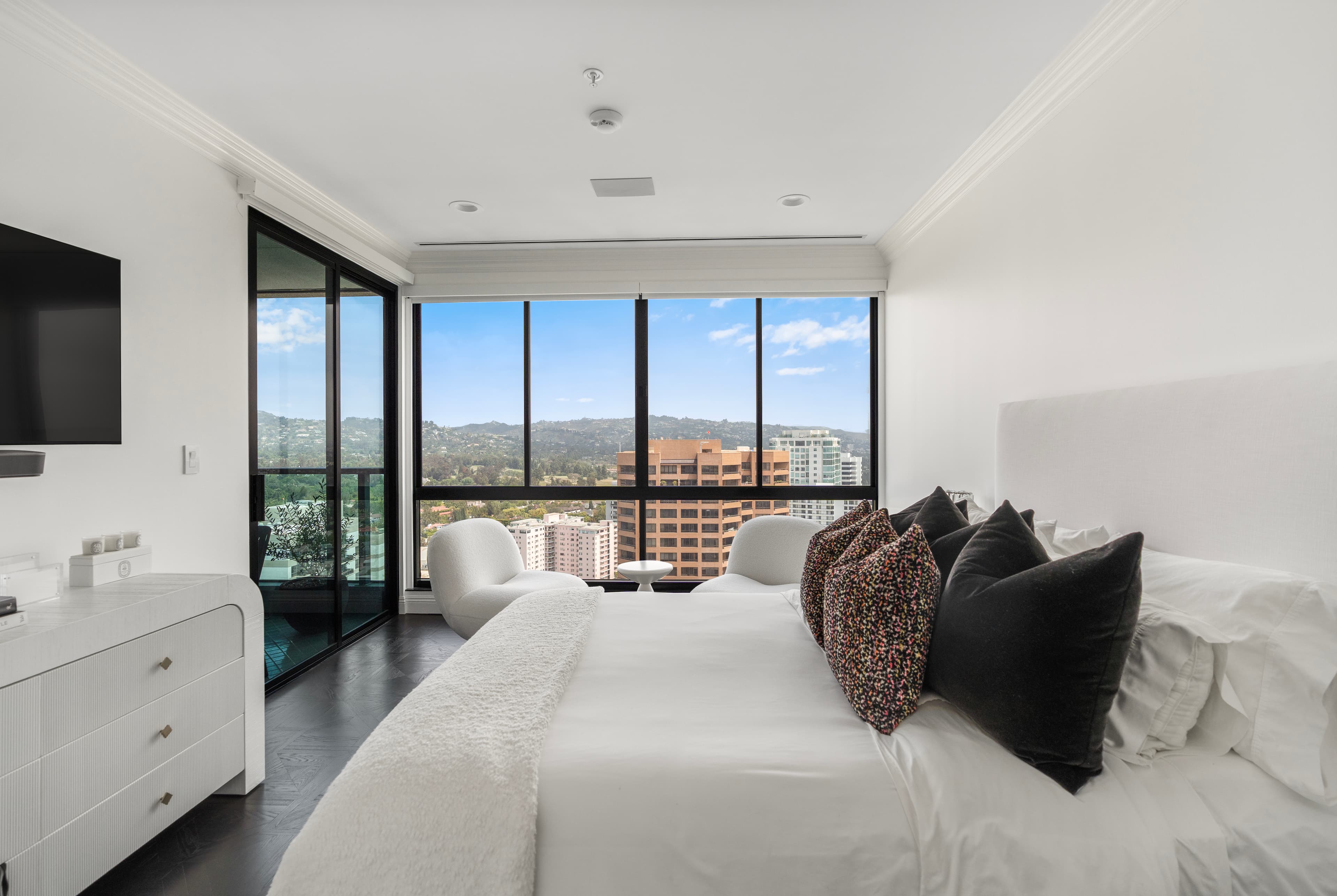 Modern white bedroom with a large bed and windows overlooking a panoramic city skyline.