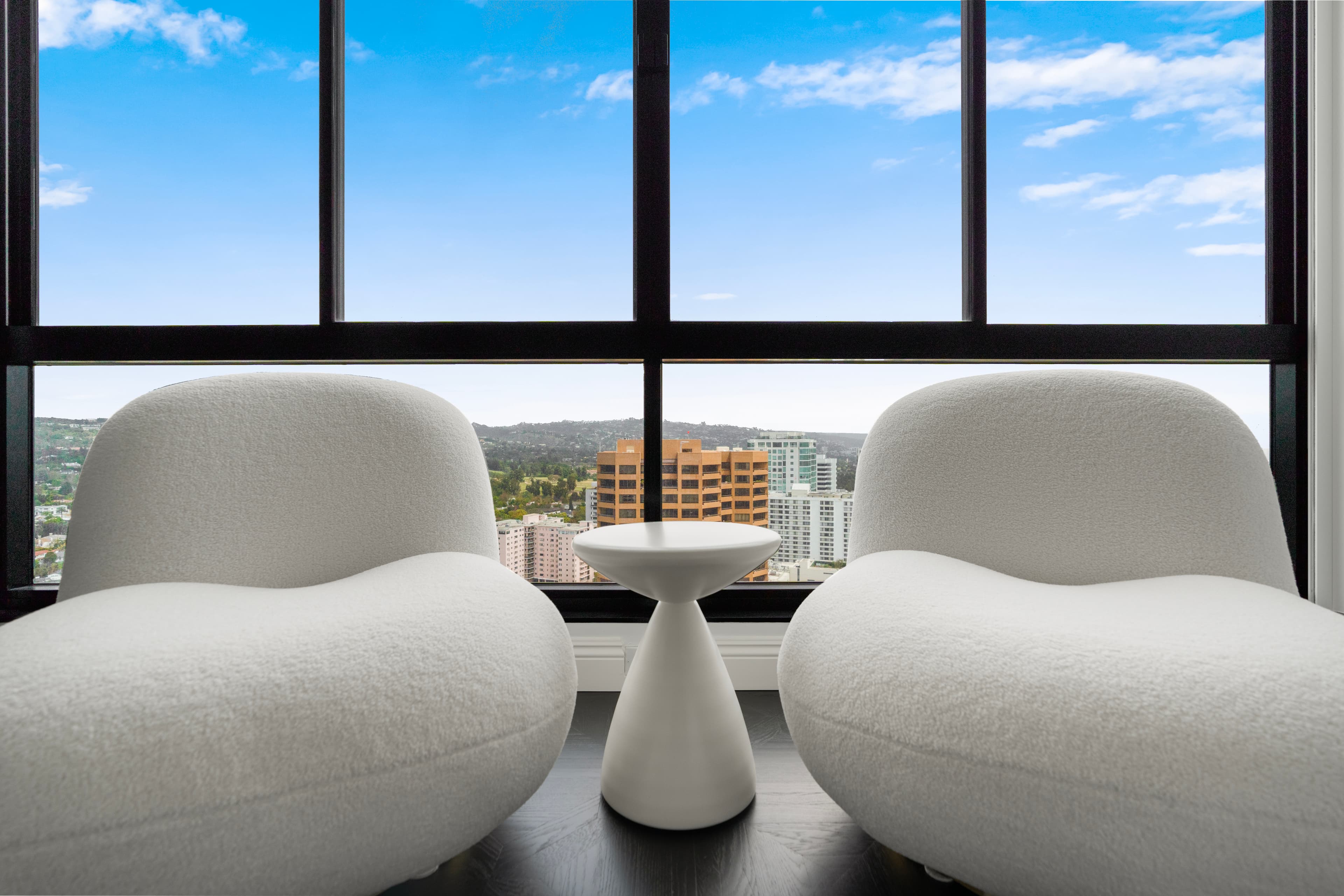 Two white textured armchairs and a pedestal table overlook a city skyline through large windows.