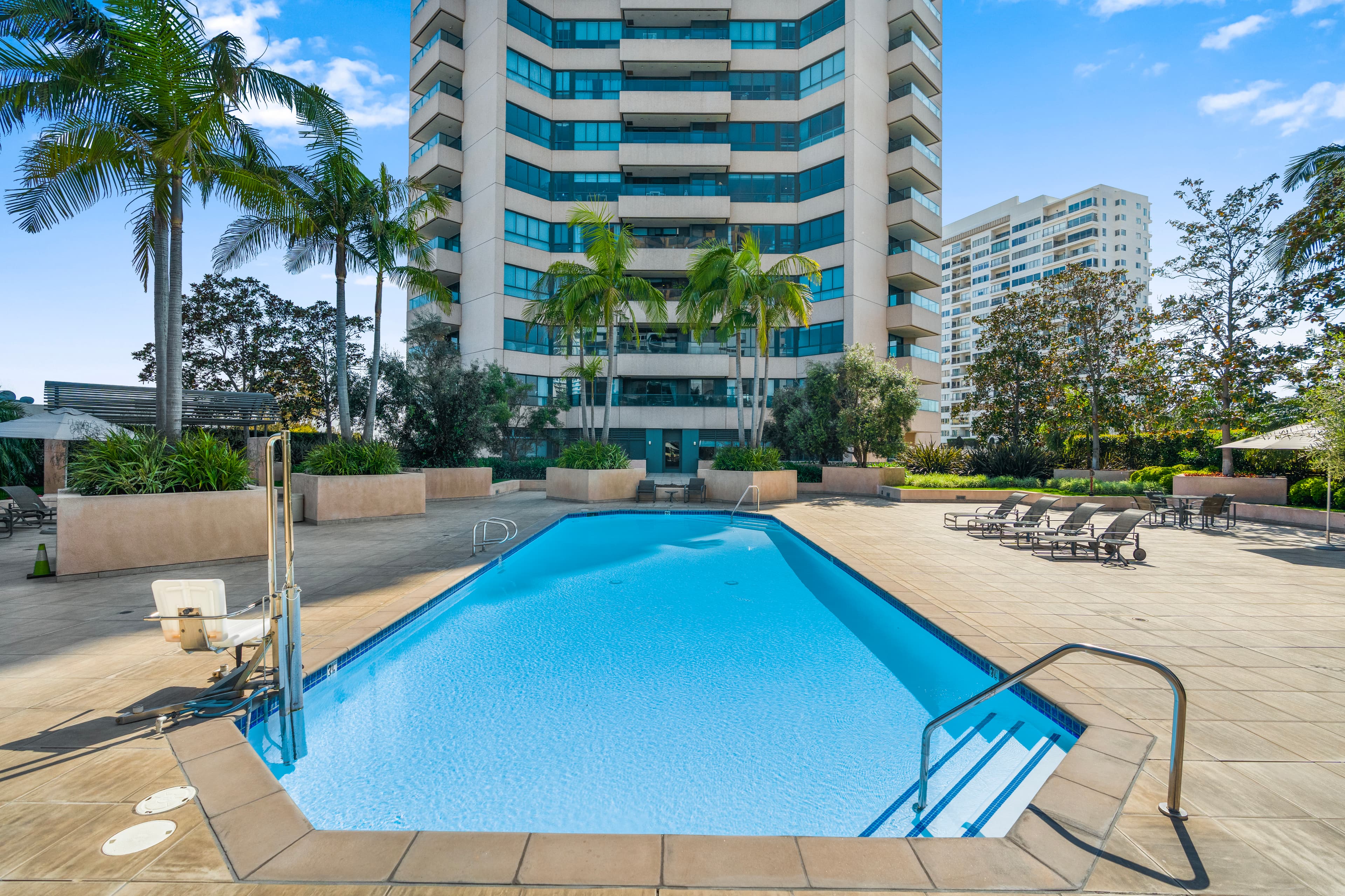 Bright blue swimming pool on a sunny patio with palm trees and a high-rise building.