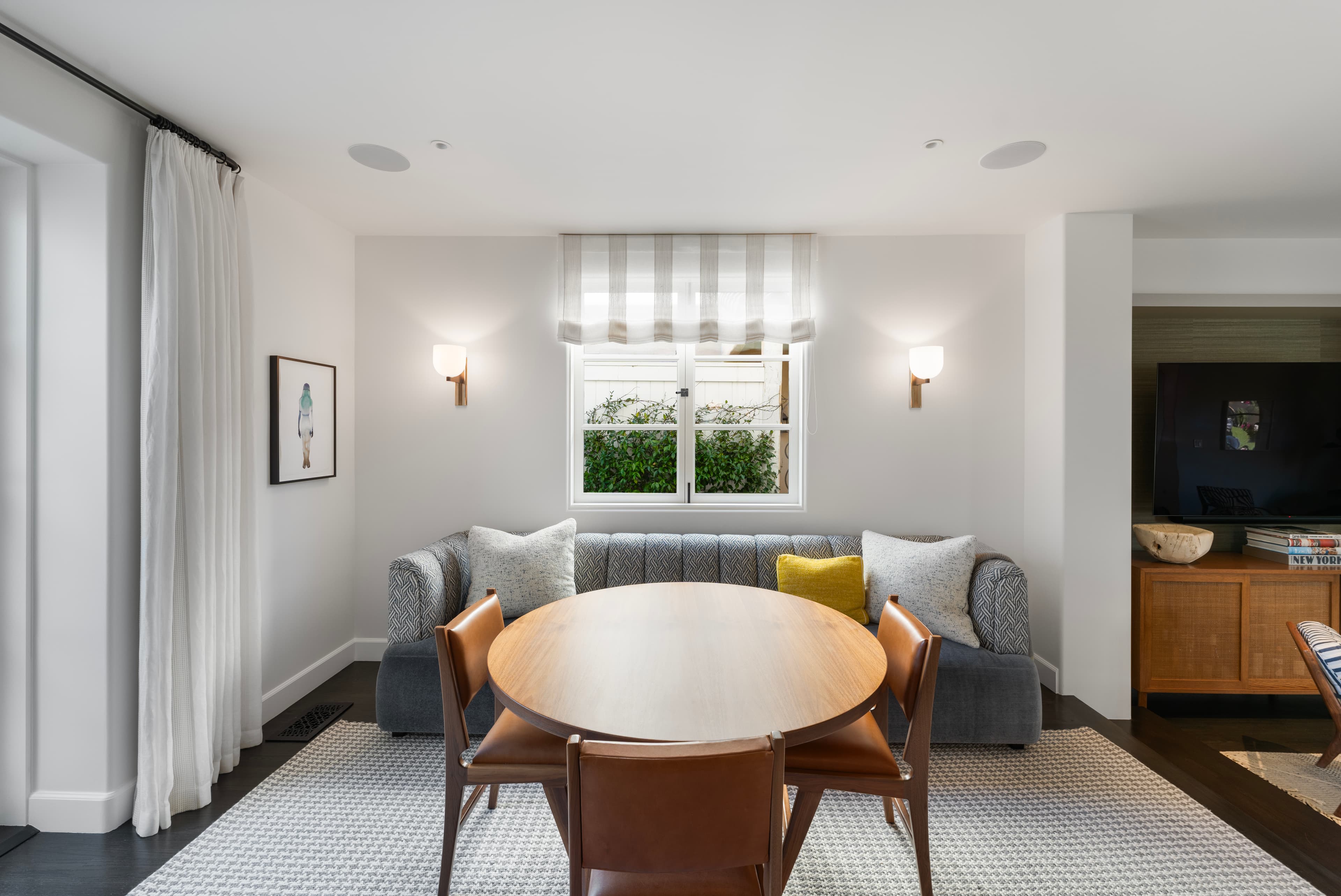 Round wooden table with leather chairs and a gray patterned banquette under a bright window.
