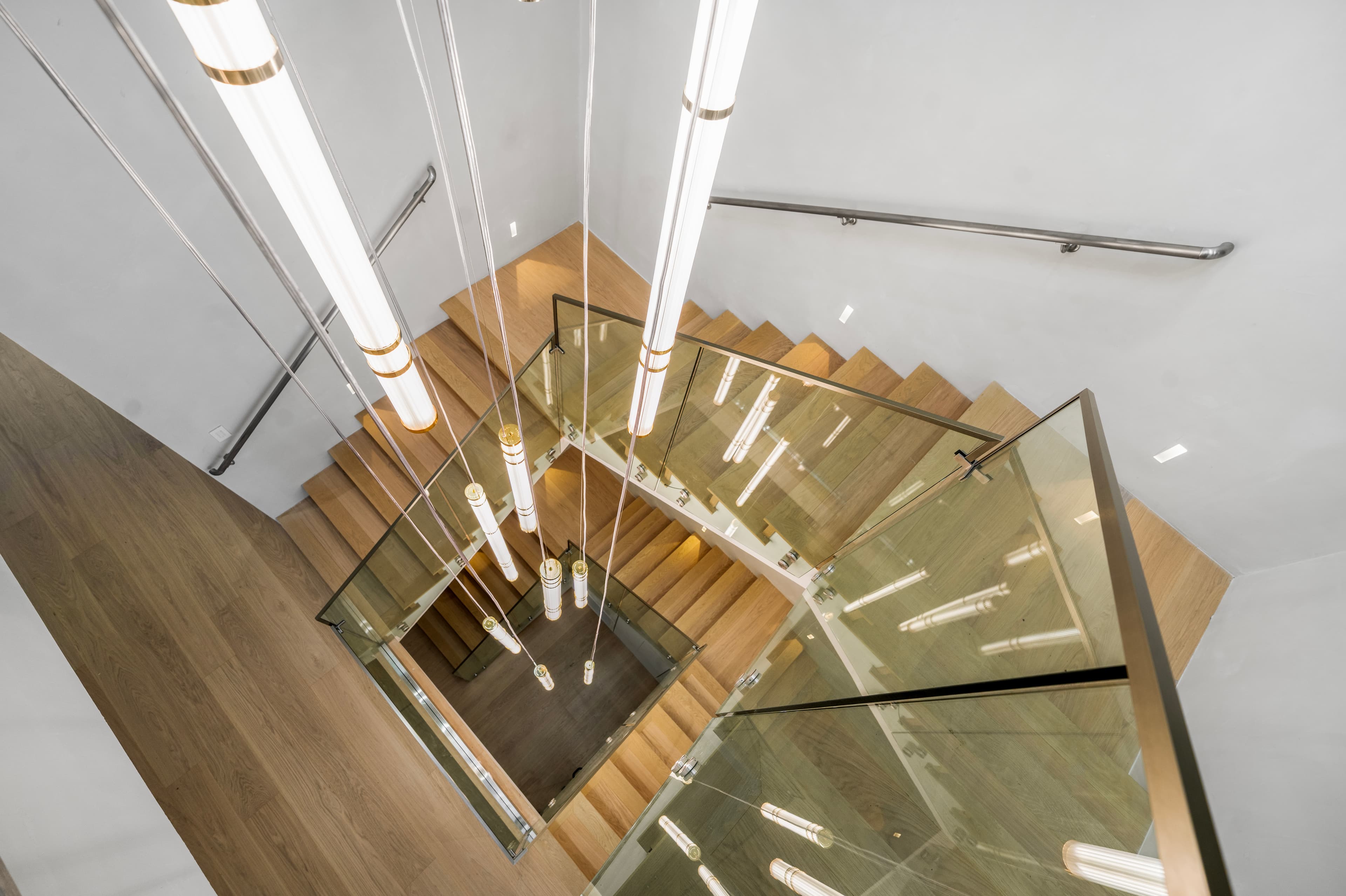 Modern wooden staircase with glass railings and long vertical tube lights viewed from above.