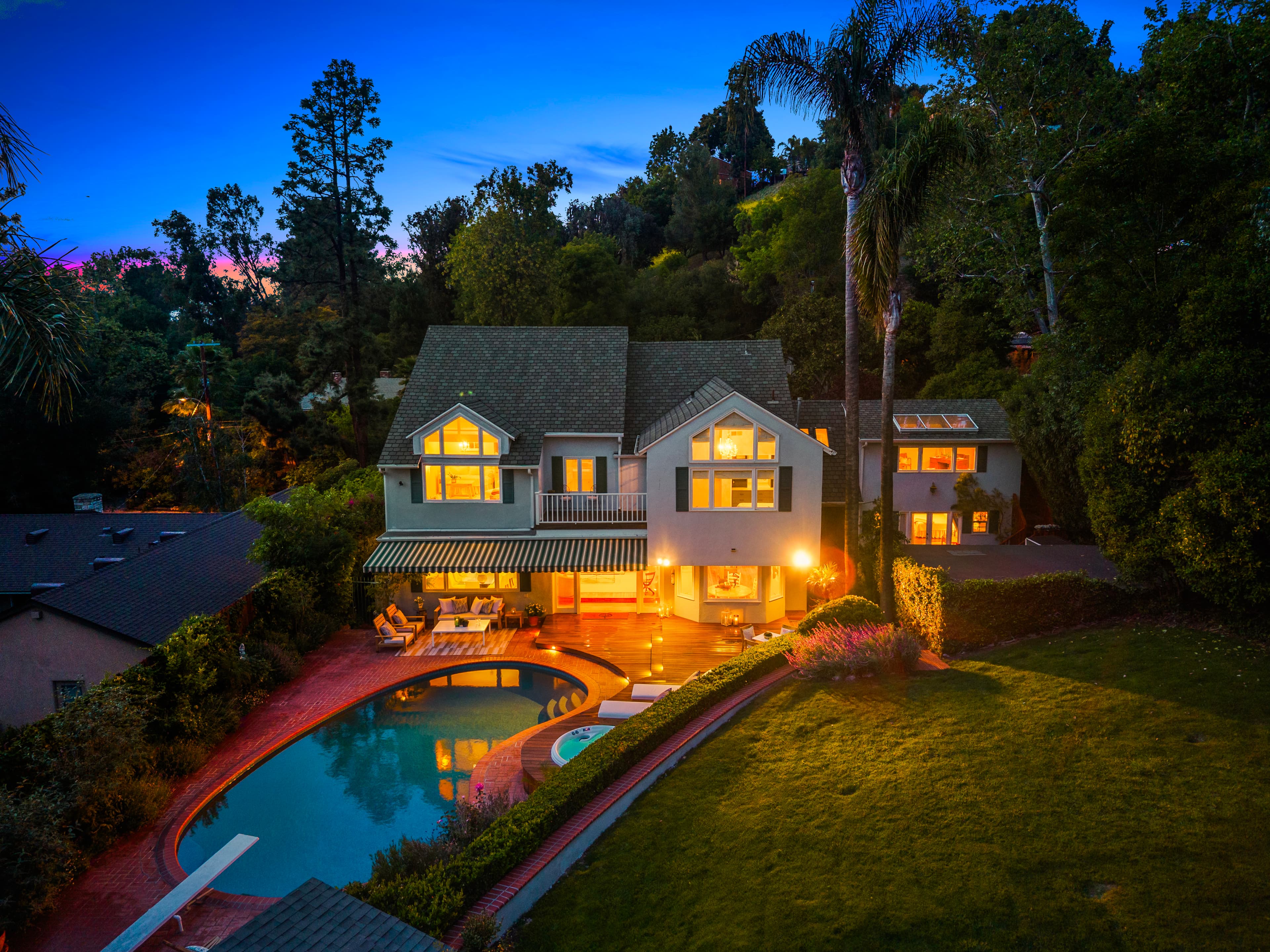 Two-story home with glowing windows, a swimming pool, and lush gardens at twilight.