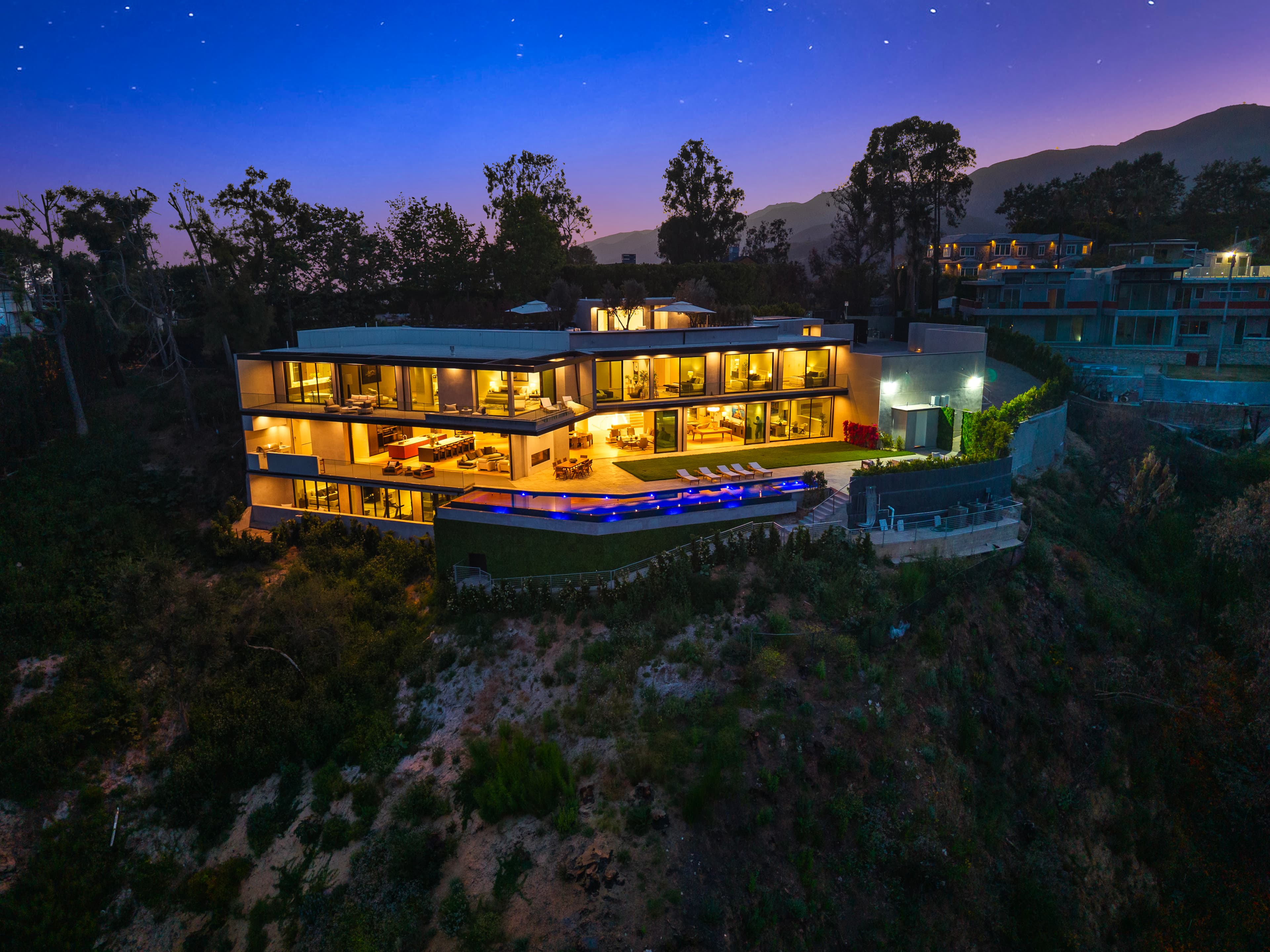 Modern hillside mansion at dusk with glowing glass walls and a blue-lit infinity pool.