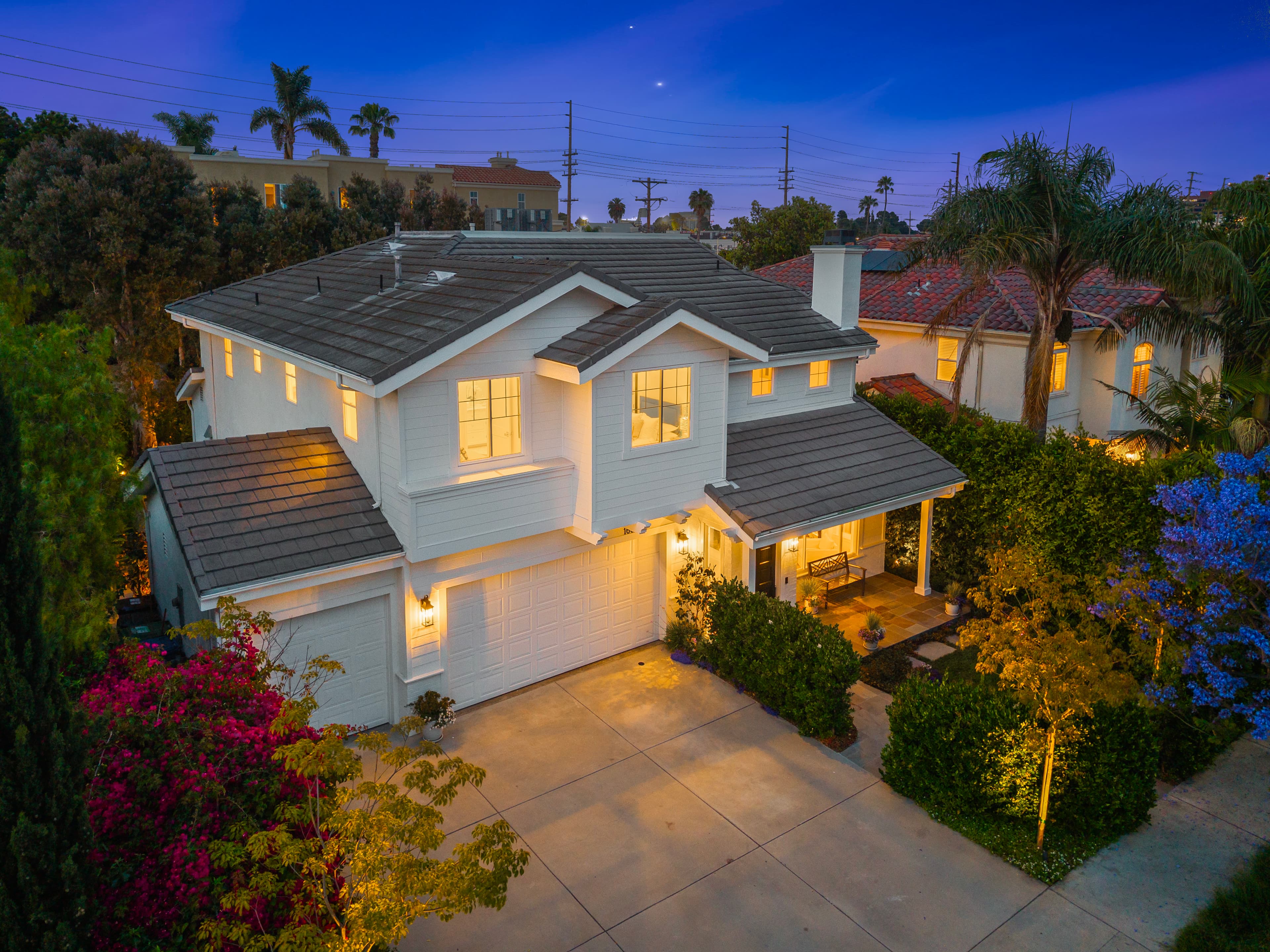 Two-story white house with glowing windows and gray roof at twilight, featuring lush landscaping.