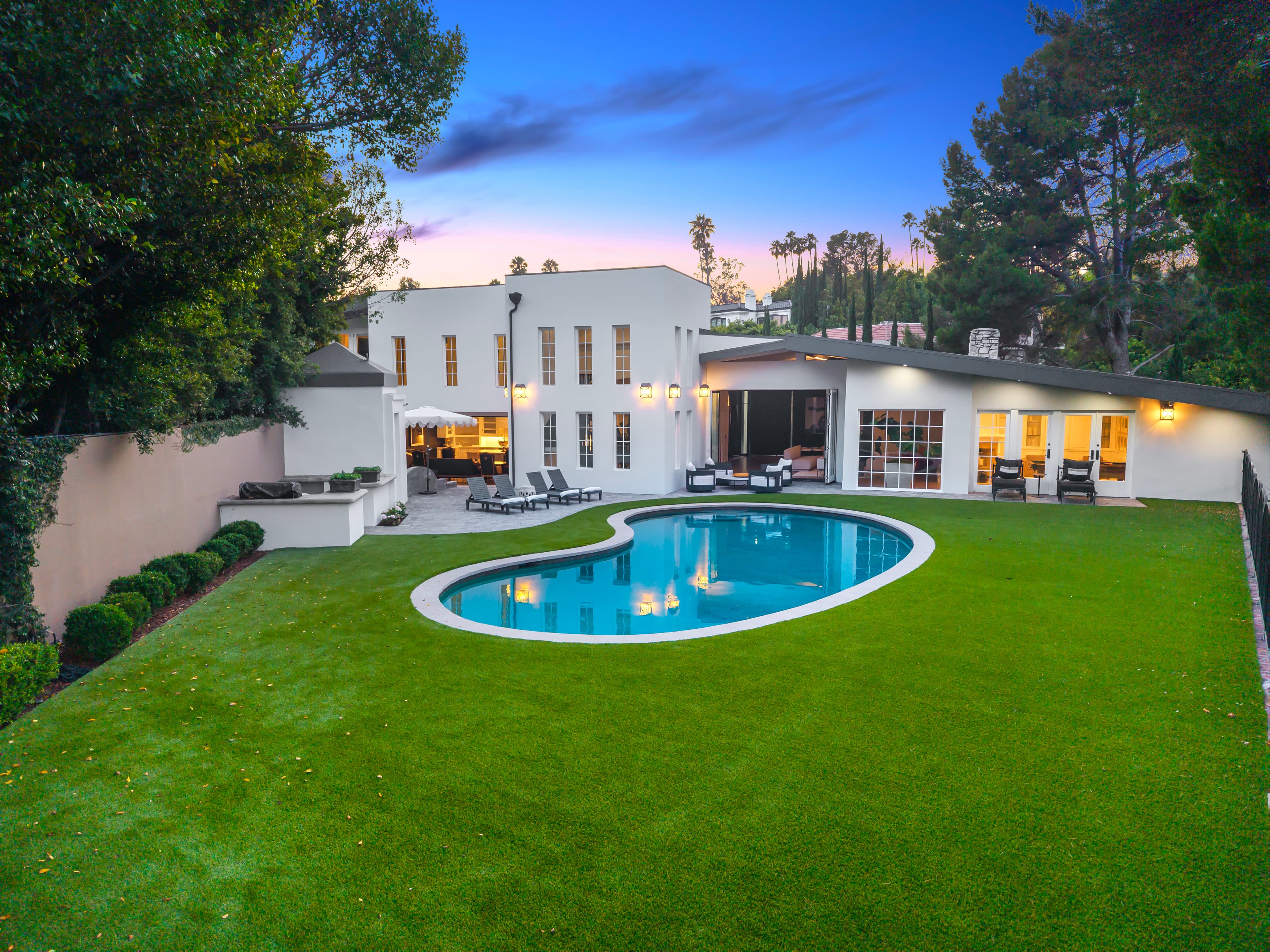 Modern white luxury home with a kidney-shaped pool and manicured lawn under a twilight sky.