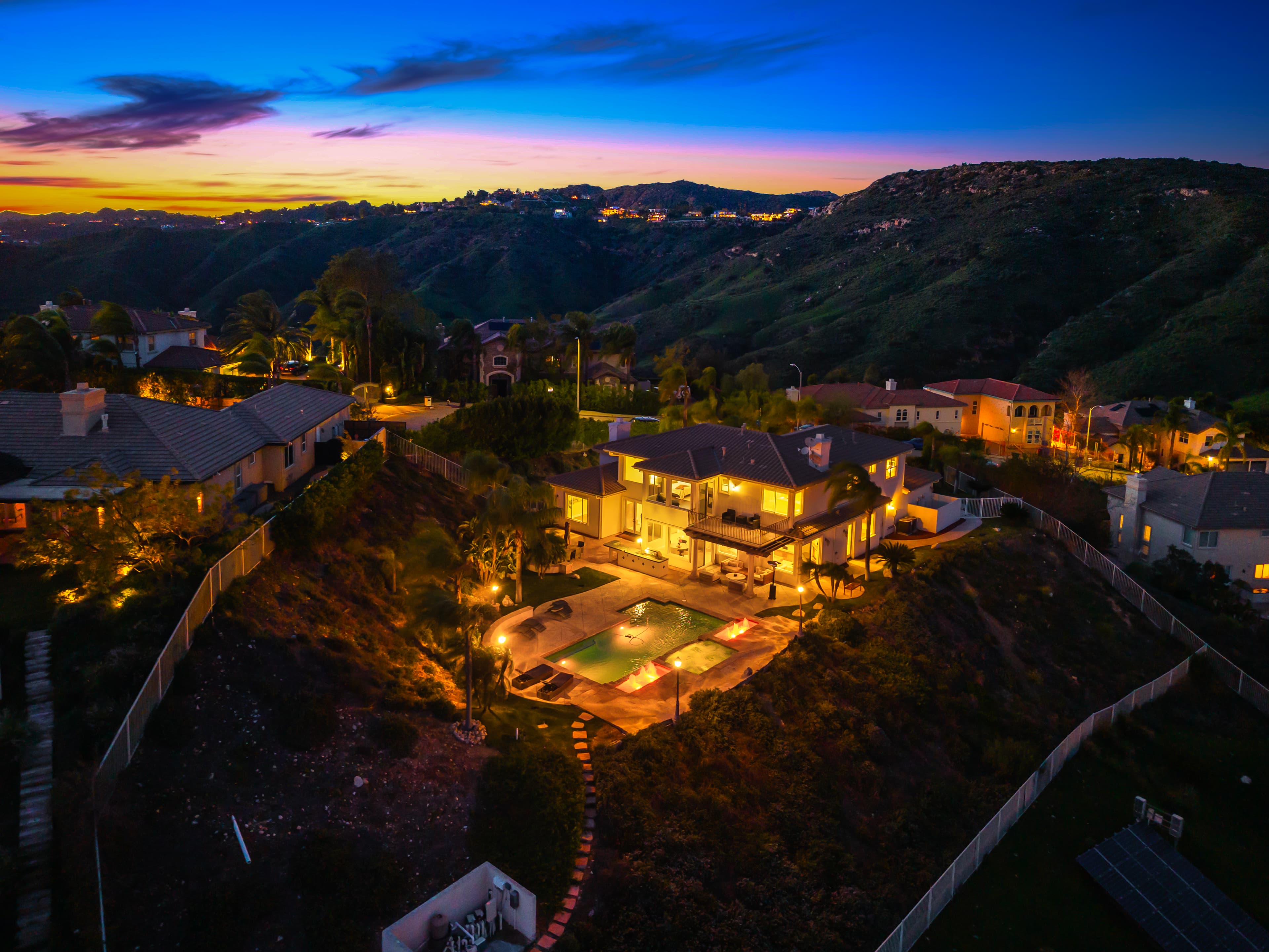 Luxury hillside home with a glowing pool and patio under a vibrant sunset sky.