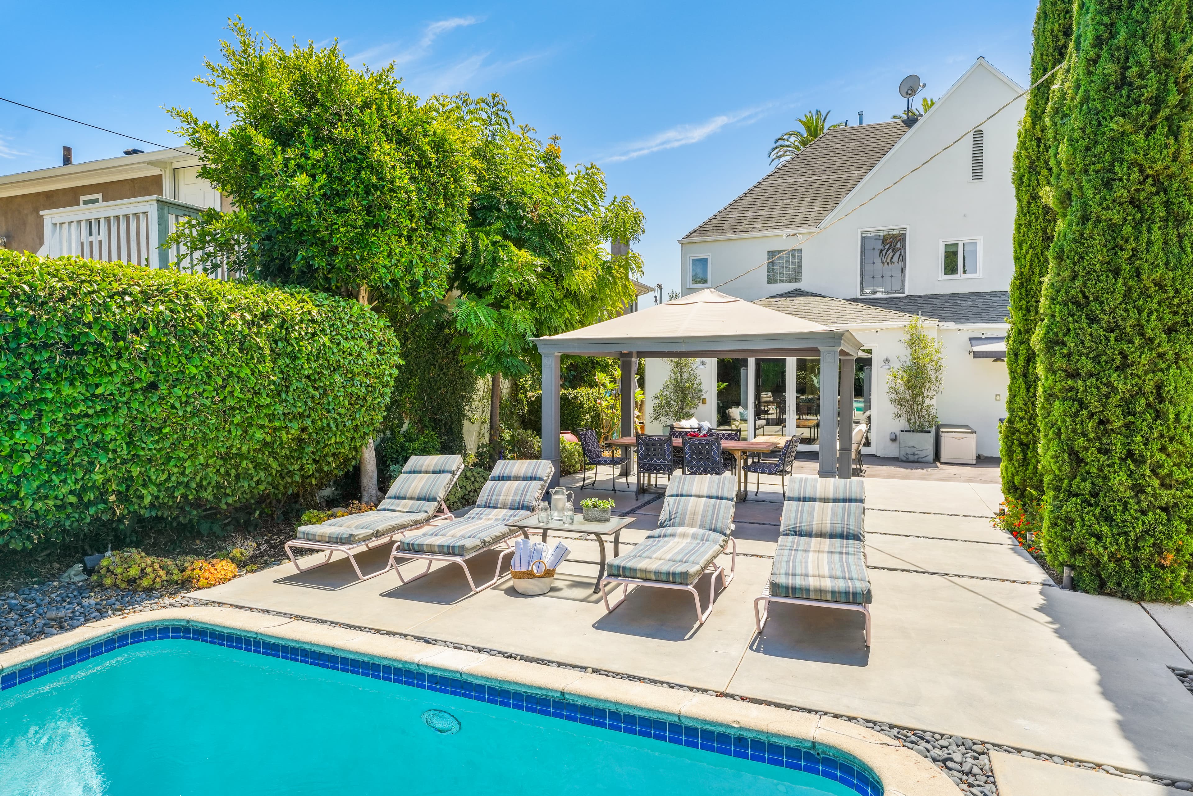 Sunny backyard with blue pool, striped lounge chairs, and gazebo next to a white house.